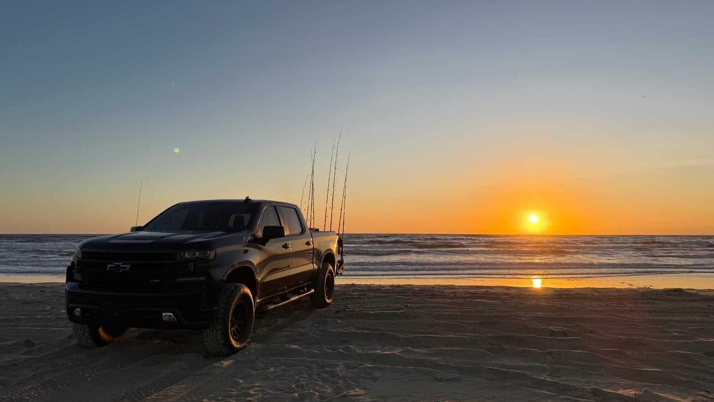 Real photo: Beach 4x4's black Silverado Trail Boss on the beach at sunrise with fishing rods in the sand.